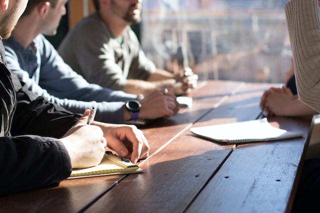 People around a wooden table, talking and making notes. Sun shines in through a nearby window.