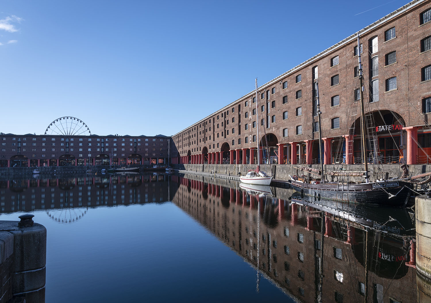 Serene looking scene of still water across the iconic Albert Dock. Traditional boats are moored up in front of the Tate Liverpool art gallery, the Wheel of Liverpool is visible over the tops of the renovated Albert Dock warehouse buildings.