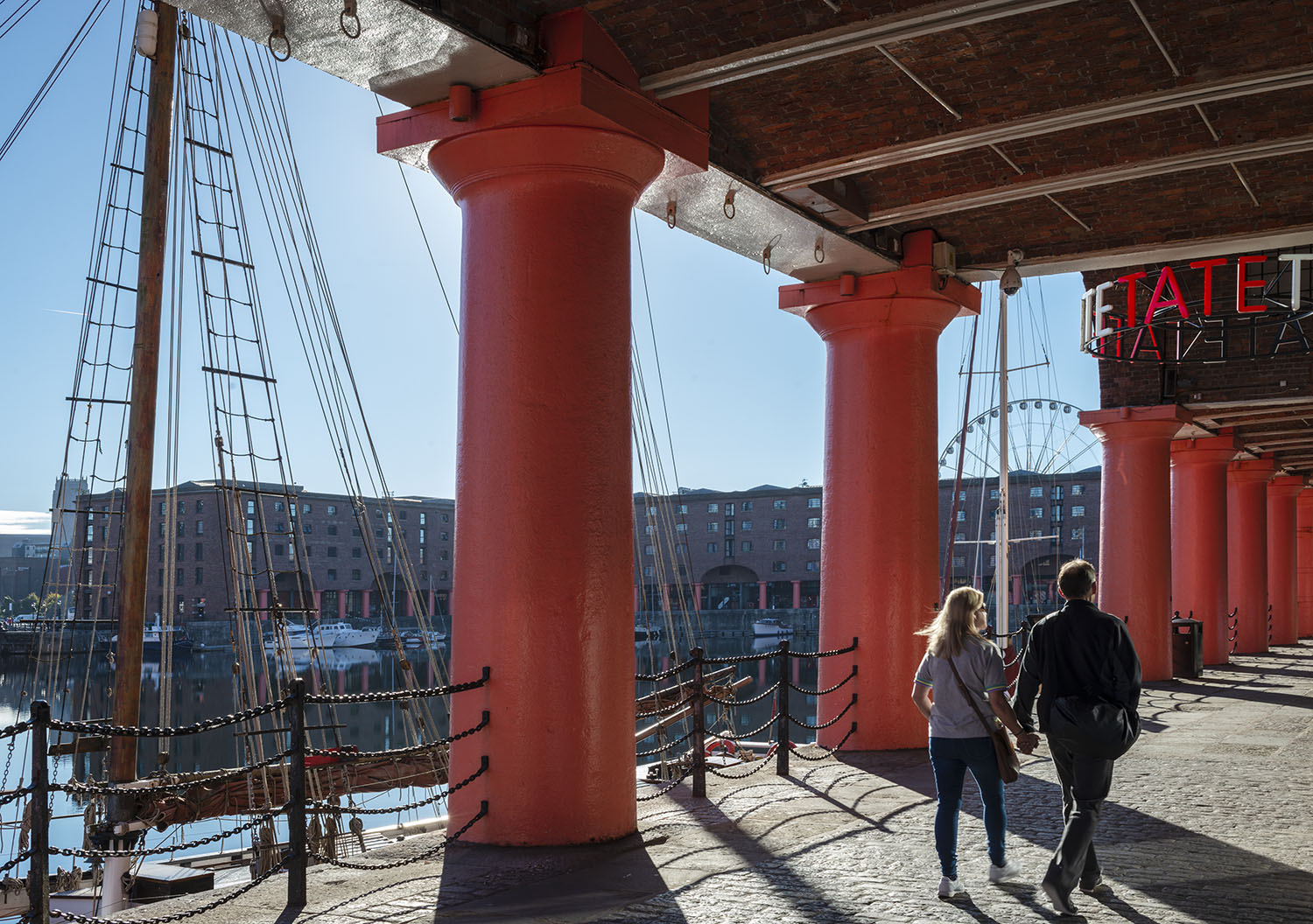 Scene looking out across the dock between the red pillars of the historical Albert Dock taken from outside Tate Liverpool art gallery. Two people walk away hand-in-hand across the foreground.