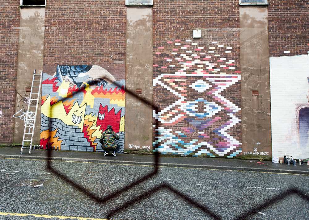 Man in camouflage jacket painting mural outside in the Baltic Triangle area of Liverpool. Shot from inside looking out through window. Man framed through hexagonal markings on window glass.