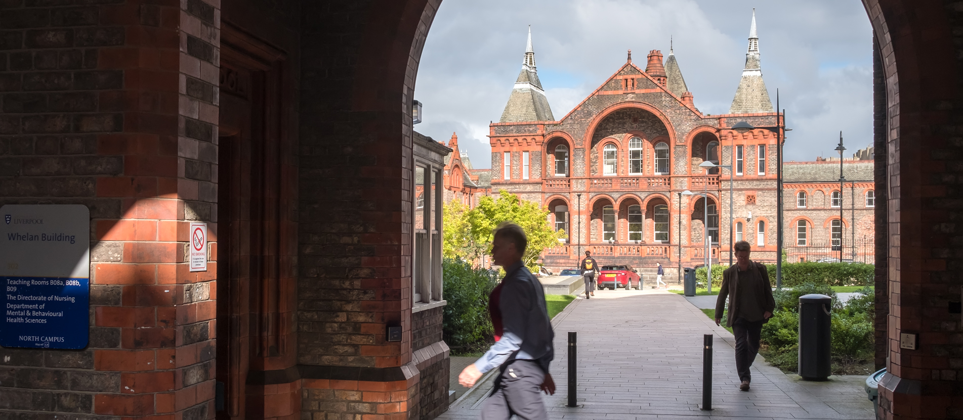 Campus exterior looking through arches of Whelan Building, various people walking are in shot, moody grey clouds are in the background.